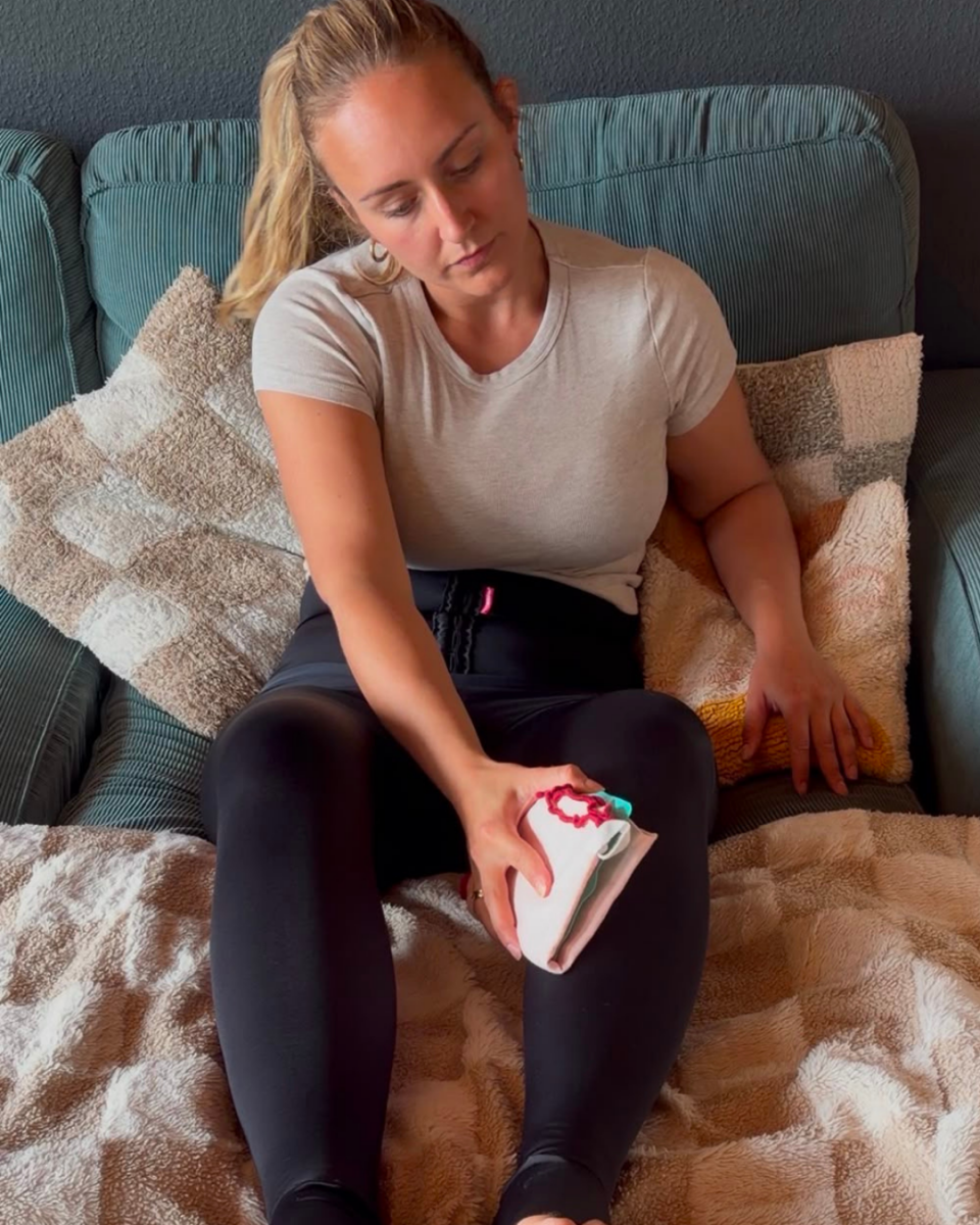 Lipödem Heldin Juliane sitzt auf dem Sofa und trägt das TBfLg variant Mieder von LIPOELASTIC. Mit einem Kühlpack kühlt sie ihre Beine vorsichtig über der Kompression.