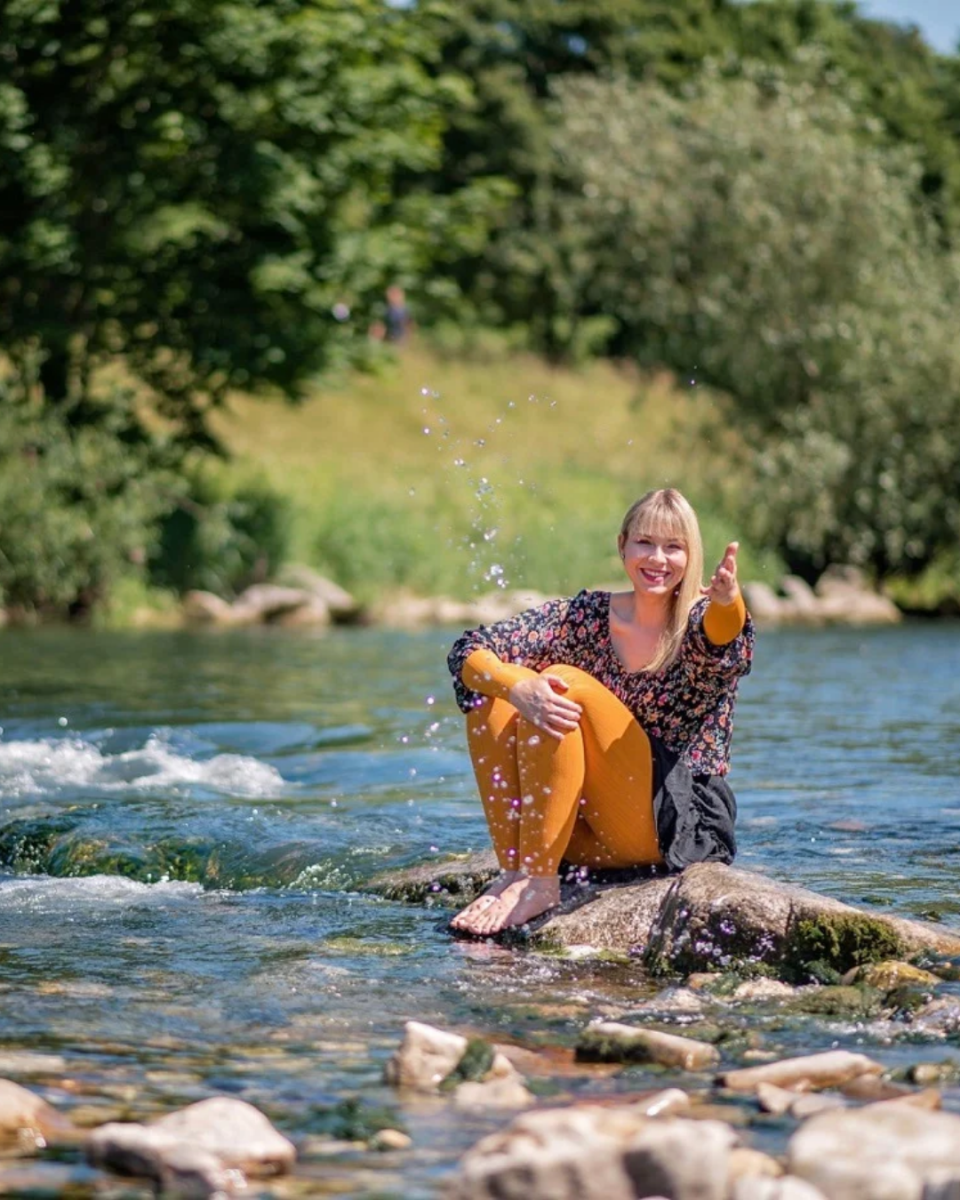 Tina Schwarz sitzt auf ein paar Steinen am Rande eines Flusses. Mit der linken Hand hat sie etwas Wasser Richtung Kamera gespritzt.